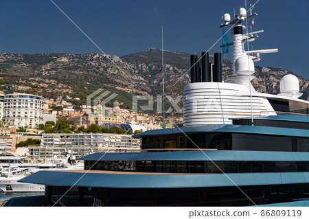 Landmarks of Monaco through decks of huge yacht of blue color in port Hercule at sunny day, glossy board of the motor boat, sun reflection on glossy board, mountains are on background 86809119