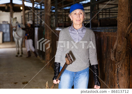 Smiling elderly woman carrying pitchfork to stack hay for horses in stable 86813712