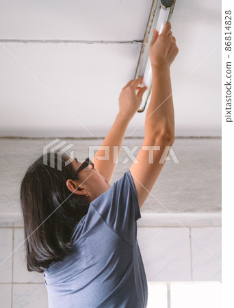 The backside of an Asian middle-aged woman changing ceiling fluorescent lamp by herself. The concept for women with men's tasks. Low angle view. The backside of an Asian middle-aged woman changing ceiling fluorescent lamp by herself. The concept for women with men's tasks. Low angle view. 86814828