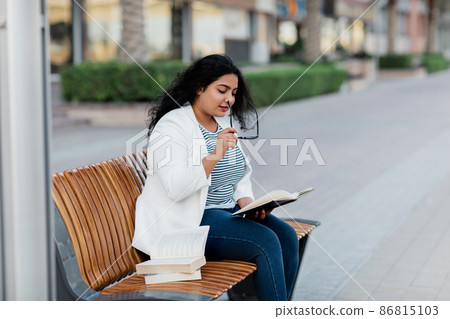 A beautiful girl is sitting on a bench and reading a book. The concept of urban lifestyle and education. Side view. 86815103