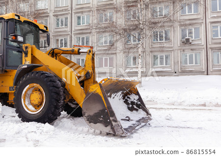 Big orange tractor cleans up snow from the road and loads it into the truck. Cleaning and cleaning of roads in the city from snow in winter. Snow removal after snowfall and blizzards. Big orange tractor cleans up snow from the road and loads it into the truck. Cleaning and cleaning of roads in the city from snow in winter. Snow removal after snowfall and blizzards. 86815554