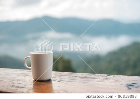White mug of hot coffee or tea on wooden table in the morning with mountain and nature background 86819888