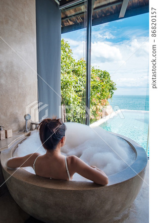 woman in white swimsuit relaxing in the bathtub with bubble foam against ocean background. Summer, relaxing and vacation concepts woman in white swimsuit relaxing in the bathtub with bubble foam against ocean background. Summer, relaxing and vacation concepts 86820157