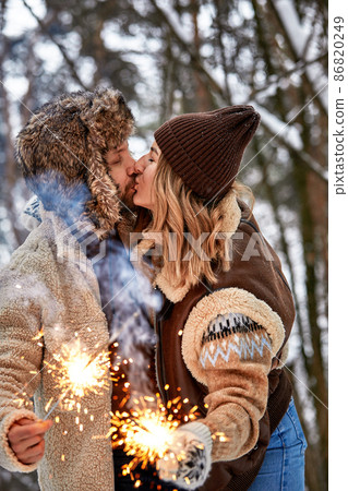 Selective focus. Couple Love Story in Snow Forest Kissing and Holding Sparklers. Couple in Winter Nature. Couple Celebrating. Valentine's day date. Selective focus. Couple Love Story in Snow Forest Kissing and Holding Sparklers. Couple in Winter Nature. Couple Celebrating. Valentine's day date. 86820249