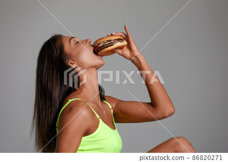 Sexy athletic woman eating hamburger on gray background Sexy athletic woman eating hamburger on gray background 86820271