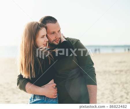 Young adult couple standing on coast and huging each other and looking away to someone. Young adult couple standing on coast and huging each other and looking away to someone. 86822482