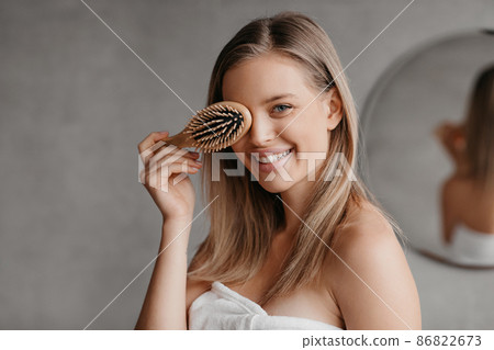 Hair care concept. Pretty young lady wrapped in white towel covering eye with hair brush, posing in bathroom interior 86822673