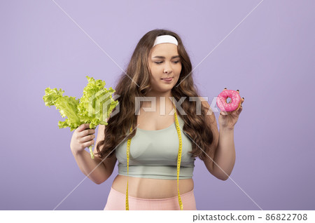 Hungry millennial european plus size lady in sportswear with measuring tape hold salad and looking at donut Hungry millennial european plus size lady in sportswear with measuring tape hold salad and looking at donut 86822708