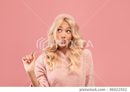 Pretty blonde woman stretching chewing gum from mouth and looking away at free space, pink studio background 86822932