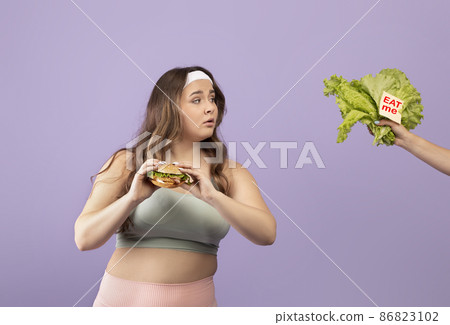 Interested hungry european young plus size lady in sportswear hold burger, look at hand with vegetables Interested hungry european young plus size lady in sportswear hold burger, look at hand with vegetables 86823102
