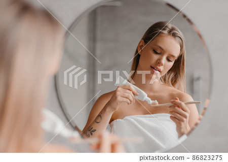 Young lady cleaning teeth in the morning, applying toothpaste on toothbrush, standing in front of mirror in bathroom 86823275