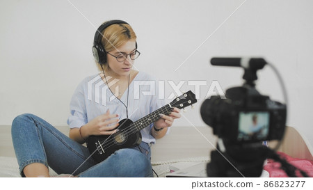 A young woman with glasses is sitting on the sofa, smiling and playing the ukulele to the camera. Playing the ukulele. Video Blogger 86823277