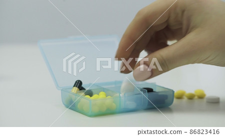 Close-up of a pillbox with different tablets. A person's hands put pills in a pillbox Close-up of a pillbox with different tablets. A person's hands put pills in a pillbox 86823416