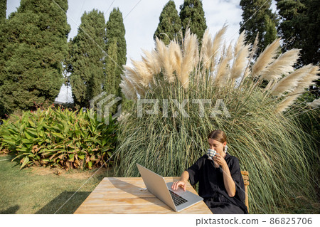 Girl use laptop and drink tea at table on lawn Girl use laptop and drink tea at table on lawn 86825706