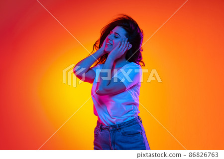 Excited young beautiful caucasian girl with long dark hair dancing isolated on orange background in neon light, filter. Concept of emotions, ads, sale Excited young beautiful caucasian girl with long dark hair dancing isolated on orange background in neon light, filter. Concept of emotions, ads, sale 86826763