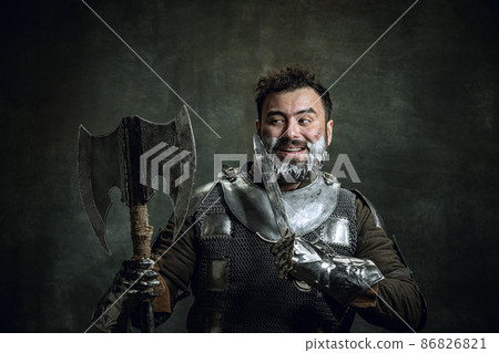 Comic portrait of medieval warrior or knight with dirty wounded face shaving with an ax isolated over dark vintage background. Comparison of eras, history, beauty Comic portrait of medieval warrior or knight with dirty wounded face shaving with an ax isolated over dark vintage background. Comparison of eras, history, beauty 86826821