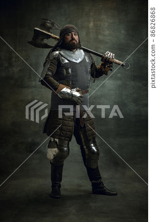 Vintage portrait of brutal seriuos man, medieval warrior or knight with wounded face in chain mail holding ax isolated over dark background. Comparison of eras 86826868