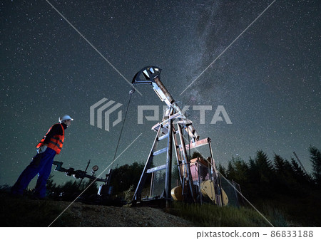 Bottom view of man in workwear standing to the left of oil pump and watching for its activities. Mechanic controls efficient night work of oil drilling rig. Starry sky and forest on background. Bottom view of man in workwear standing to the left of oil pump and watching for its activities. Mechanic controls efficient night work of oil drilling rig. Starry sky and forest on background. 86833188