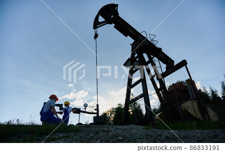 Oil industry equipment. On backdrop of blue sky and oil well pump jack adult engineer in protective construction helmet checking pipeline with kid boy. They interesting spending time in borehole. Oil industry equipment. On backdrop of blue sky and oil well pump jack adult engineer in protective construction helmet checking pipeline with kid boy. They interesting spending time in borehole. 86833191