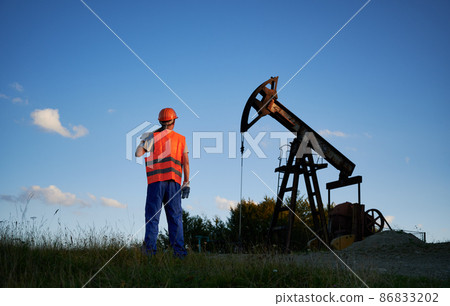 Rear view of oilman in orange uniform with working tool who watching at pump oil working in blue sky background. Petroleum extraction machine in energetic industry. 86833202