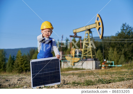 Cute child in construction helmet holding portable solar panel and giving thumbs up. Male kid showing approval gesture while standing on territory of oil field with pump jack on blurred background. 86833205