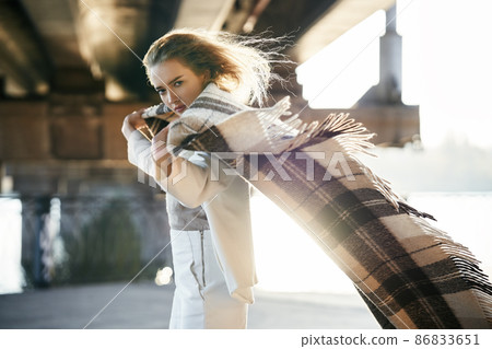 Beautiful young woman with blanket looking to camera on windy sunny day posing on urban street background Beautiful young woman with blanket looking to camera on windy sunny day posing on urban street background 86833651