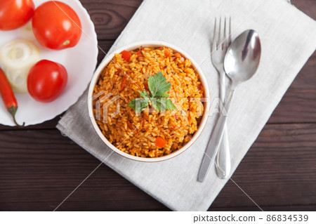 Jollof rice, tomatoes and hot peppers on a blue plate, fork, spoon on a linen napkin on a brown wooden background. National cuisine of Africa. 86834539