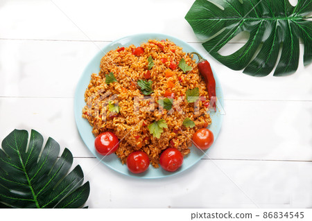 Jollof rice with tomatoes and hot peppers on a blue plate on a white plate with monstera palm leaves. National cuisine of Africa. 86834545