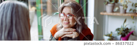 Grey haired woman with glasses looks at friend sitting at small table in cozy cafe 86837022