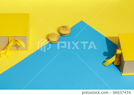 Yellow spring gift box with ribbon bow and two french macaroons over yellow and blue background. Greeting for International Womens Day on March 8, mothers day or spring concept. Copy space Yellow spring gift box with ribbon bow and two french macaroons over yellow and blue background. Greeting for International Womens Day on March 8, mothers day or spring concept. Copy space 86837434