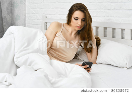 Young sleepy beautiful woman using smartphone after awakening in morning while being in bed Young sleepy beautiful woman using smartphone after awakening in morning while being in bed 86838972