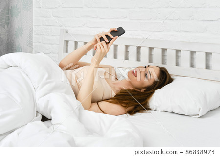 Young smiling woman in bed with smartphone scrolling through social media after waking up in morning Young smiling woman in bed with smartphone scrolling through social media after waking up in morning 86838973