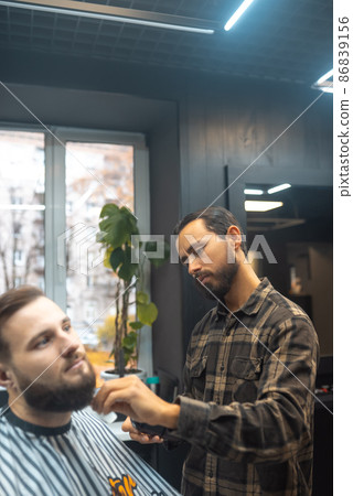 Barber works with a beard clipper. Hipster client getting haircut. 86839156