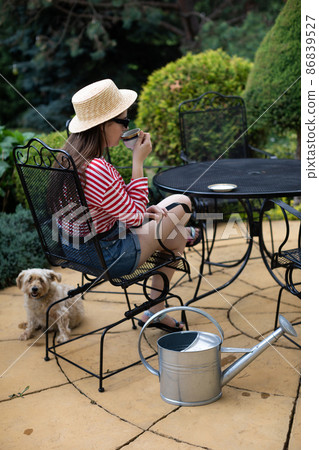 A shaggy dog sits on the terrace and a girl sits on a chair and drinks coffee from a cup. A shaggy dog sits on the terrace and a girl sits on a chair and drinks coffee from a cup. 86839527