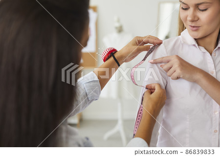 Close up of seamstress or tailor work with woman client in fashion atelier or style studio. Designer or dressmaker take customer measures with tape create clothes or garment in workshop. 86839833