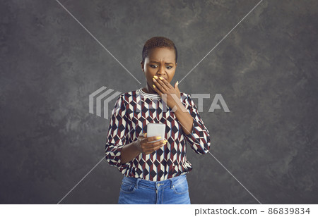 Portrait of an anxious frightened young African American woman looking at the phone in her hands. Woman scared to see bad news or photos. Gray background. Concept of negative emotions and reaction. 86839834