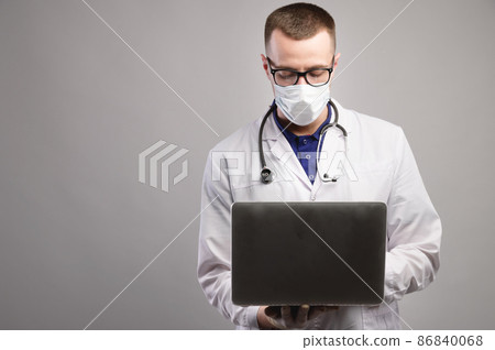 young caucasian doctor in a mask and glasses uses his laptop. studio portrait 86840068