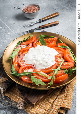 Italian cheese stracciatella burrata on plate with fresh tomatoes and arugula. Gray background. Top view Italian cheese stracciatella burrata on plate with fresh tomatoes and arugula. Gray background. Top view 86841004