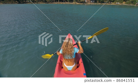 Seductive blonde woman in blue swimsuit sails on pink plastic kayak along azure sea at exotic resort backside upper above view. 86841551