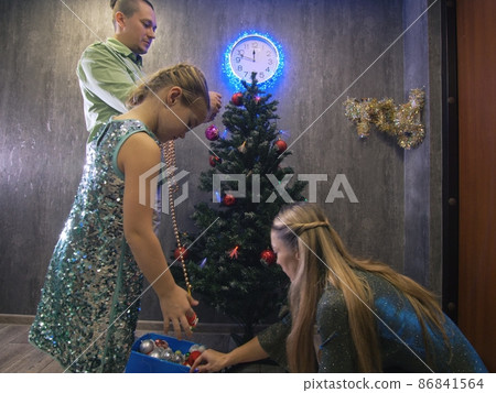The family fast and fanny decorates the Christmas tree. Side view. Time on the clock is 23.45 on wall. The family fast and fanny decorates the Christmas tree. Side view. Time on the clock is 23.45 on wall. 86841564