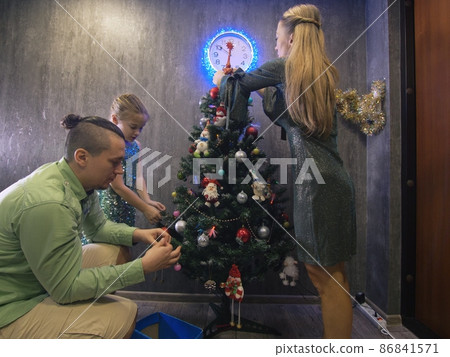 The family fast and fanny decorates the Christmas tree. Side view. Time on the clock is 23.45 on wall. The family fast and fanny decorates the Christmas tree. Side view. Time on the clock is 23.45 on wall. 86841571
