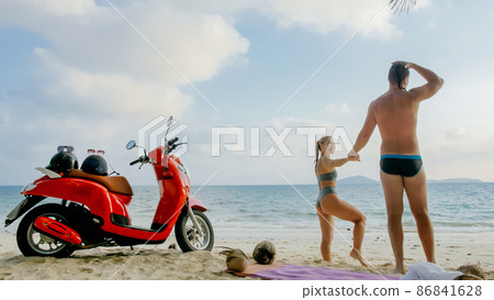 Scooter road trip. Lovely couple on red motorbike in white clothes on sand beach. People walking near the tropical palm trees, sea. Motorcycle rent. 86841628