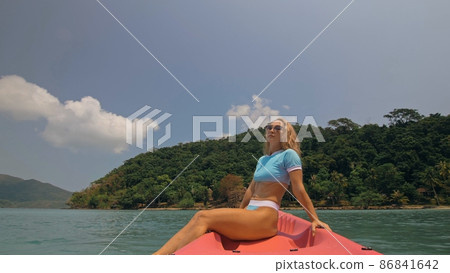 Pretty young blonde woman in stylish swimsuit and sunglasses sits on pink canoe sailing along sea against hills. Pretty young blonde woman in stylish swimsuit and sunglasses sits on pink canoe sailing along sea against hills. 86841642
