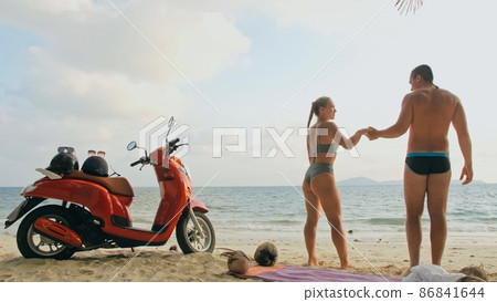 Scooter road trip. Lovely couple on red motorbike in white clothes on sand beach. People walking near the tropical palm trees, sea. Motorcycle rent. Scooter road trip. Lovely couple on red motorbike in white clothes on sand beach. People walking near the tropical palm trees, sea. Motorcycle rent. 86841644