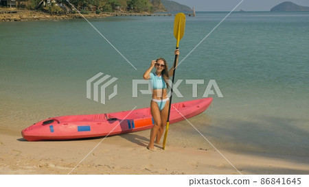 Pretty young sportswoman with sunglasses and swimsuit holds paddle posing near pink plastic canoe on sand beach at resort. Pretty young sportswoman with sunglasses and swimsuit holds paddle posing near pink plastic canoe on sand beach at resort. 86841645
