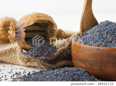 Low angle close-up of broken dry poppy seed pod with ripe poppies. White wooden table with wooden bowl full of poppy seeds. Low angle close-up of broken dry poppy seed pod with ripe poppies. White wooden table with wooden bowl full of poppy seeds. 86842119