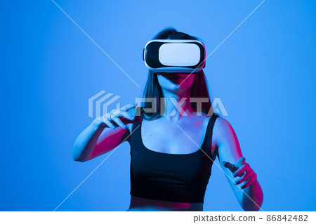 Young brunette girl in black t-shirt playing online games with a friends using VR glasses. Enjoying virtual reality headset while gaming with a friends in metaverse in neon light. 86842482