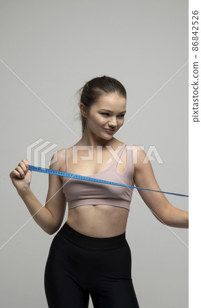 Happy attractive brunette woman with a perfect sexy body in beige top sportswear holding a blue measuring tape. Weight loss and diet concepts. Health care and healthy nutrition. Perfect slim body. 86842526