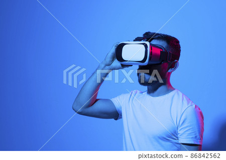 Closeup of young man wearing virtual reality goggles in modern studio with a neon light. Smartphone using with VR headset. Future technology concept. Closeup of young man wearing virtual reality goggles in modern studio with a neon light. Smartphone using with VR headset. Future technology concept. 86842562