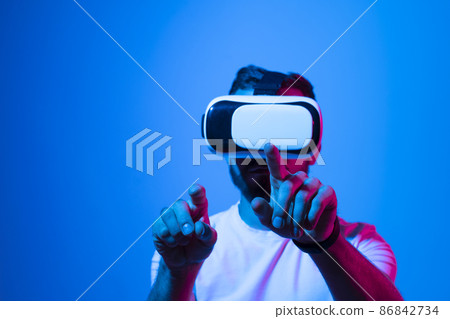 Bearded man playing online VR video game in metaverse with a friends with virtual reality goggles and trying to touch something with both hands. Future technology concept. Bearded man playing online VR video game in metaverse with a friends with virtual reality goggles and trying to touch something with both hands. Future technology concept. 86842734